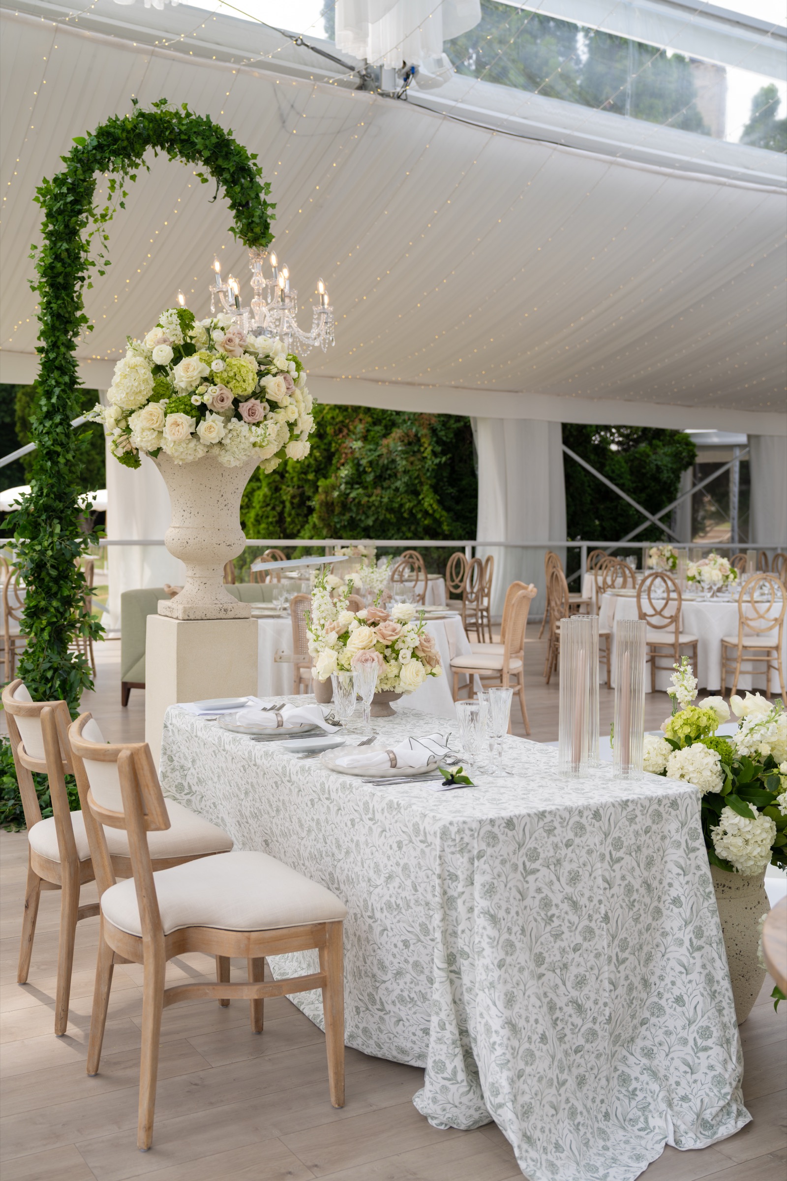 A long table in green and white toile under a topiary arch, with blush and white florals in a stone urn